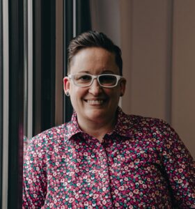 Beth, a white female presenting person with short brown hair, glasses, and a red and pink colorful shirt 