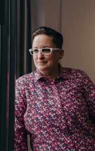 A white, female presenting person with short brown hair, glasses, and a colorful red and pink shirt looks pensively at the camera.